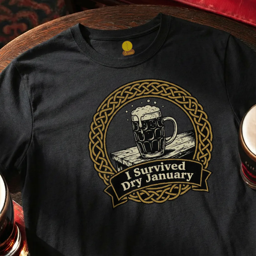Black t-shirt with a graphic of a mug of beer and 'I Survived Dry January' text, placed on a wooden surface.