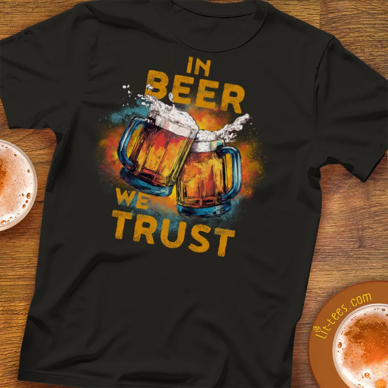 Black t-shirt with 'In Beer We Trust' graphic on a wooden surface with beer glasses.