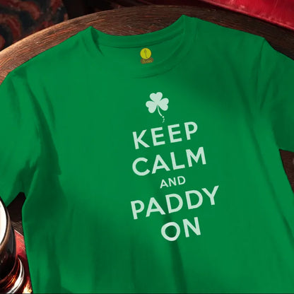 Green t-shirt with 'Keep Calm and Paddy On' text and shamrock design on a wooden surface.