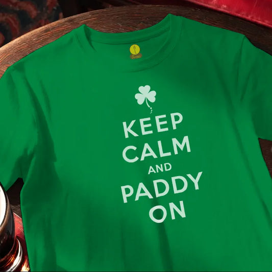 Green t-shirt with 'Keep Calm and Paddy On' text and shamrock design on a wooden surface.