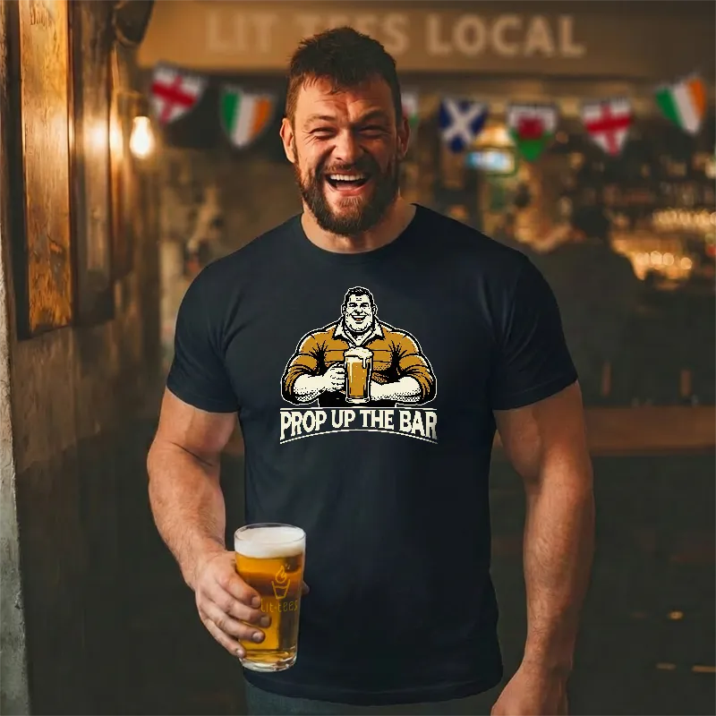 Man wearing a black t-shirt with a graphic and text saying 'Prop up the Bar', holding a beer in a bar setting.