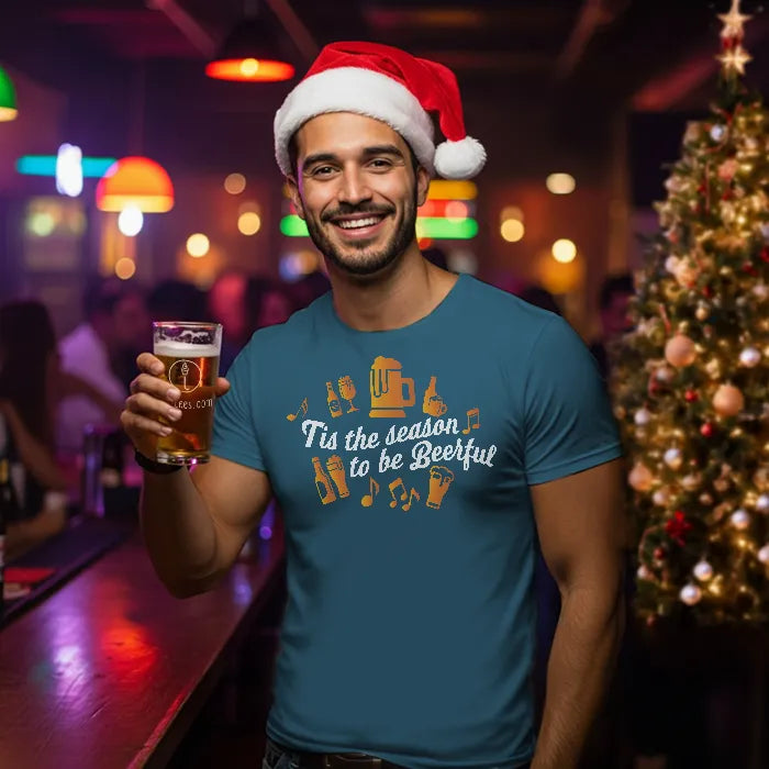 Man wearing a Santa hat and blue Christmas t-shirt reading 'Tis the season to be beerful', holding a beer in a bar setting. Glass has lit-tees.com printed on it.