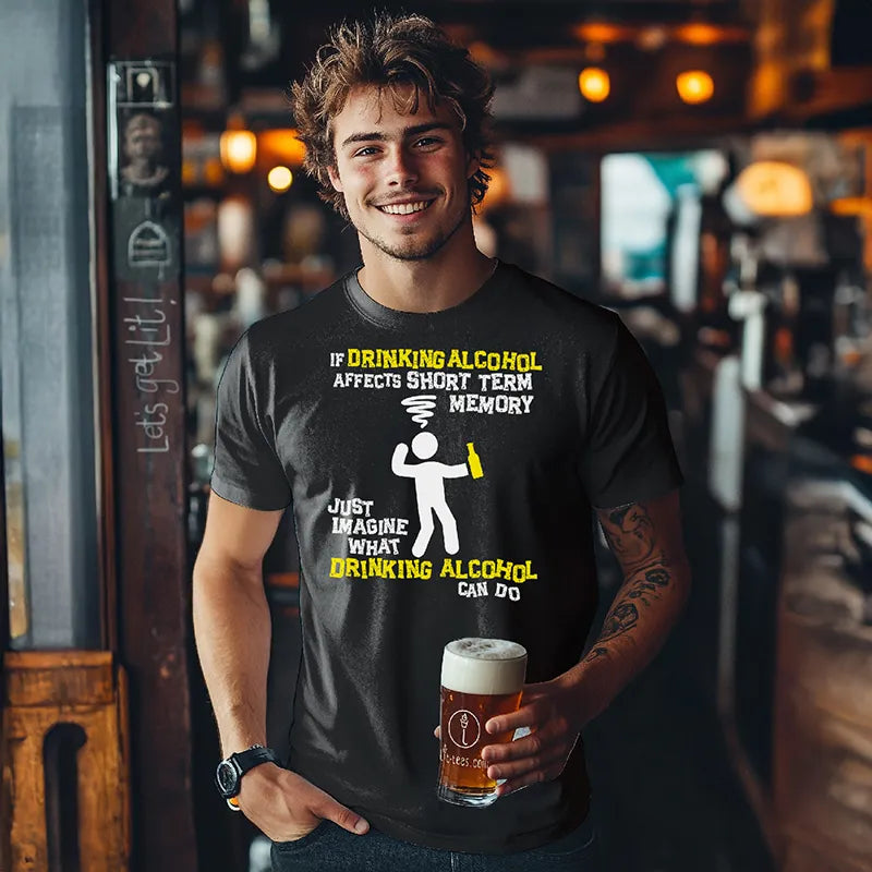 Young man holding a beer in a bar wearing a black tee shirt with a graphic character and humorous text saying 'If drinking alcohol affects short term memory just imagine what drinking alcohol can do' 